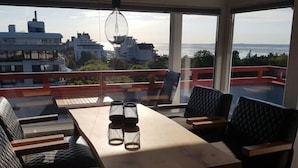 A balcony with a table and chairs, a view of buildings and the sea, and a hanging light fixture. - Little Skysuite W 94 (Cuxhaven)