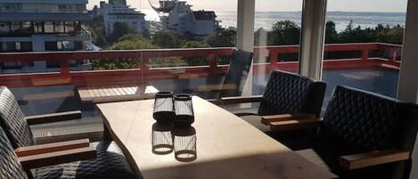 Un balcón con mesa y sillas, vista a edificios y al mar, y una lámpara colgante.