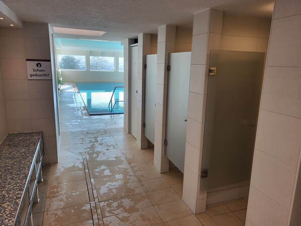 Pasillo de hotel con piso mojado, un cartel que dice “Schon geduscht?”, una zona de piscina visible a través de puertas de vidrio y un área de duchas con azulejos blancos.