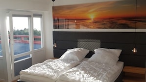 A bedroom with a bed, a large painting of a sunset, and a view of the outside. - Little Skysuite W 94 (Cuxhaven)