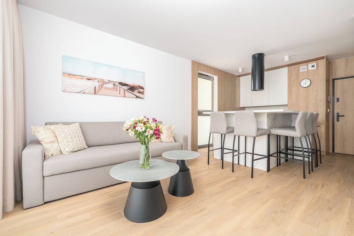 Una sala de estar moderna con un sofá, dos mesas, un jarrón de flores y una zona de cocina con taburetes de bar.