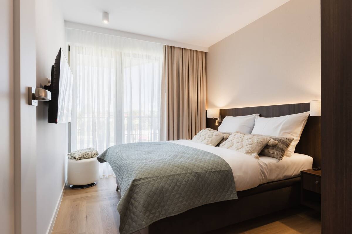 Habitación de hotel moderna con una cama grande, una mesita de noche, una lámpara, un otoman redondito y cortinas traslúcidas.