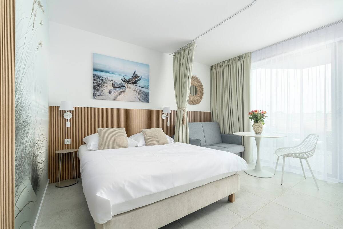 Una habitación de hotel moderna con una cama grande, una mesita redonda pequeña, un sofá gris y una silla blanca.