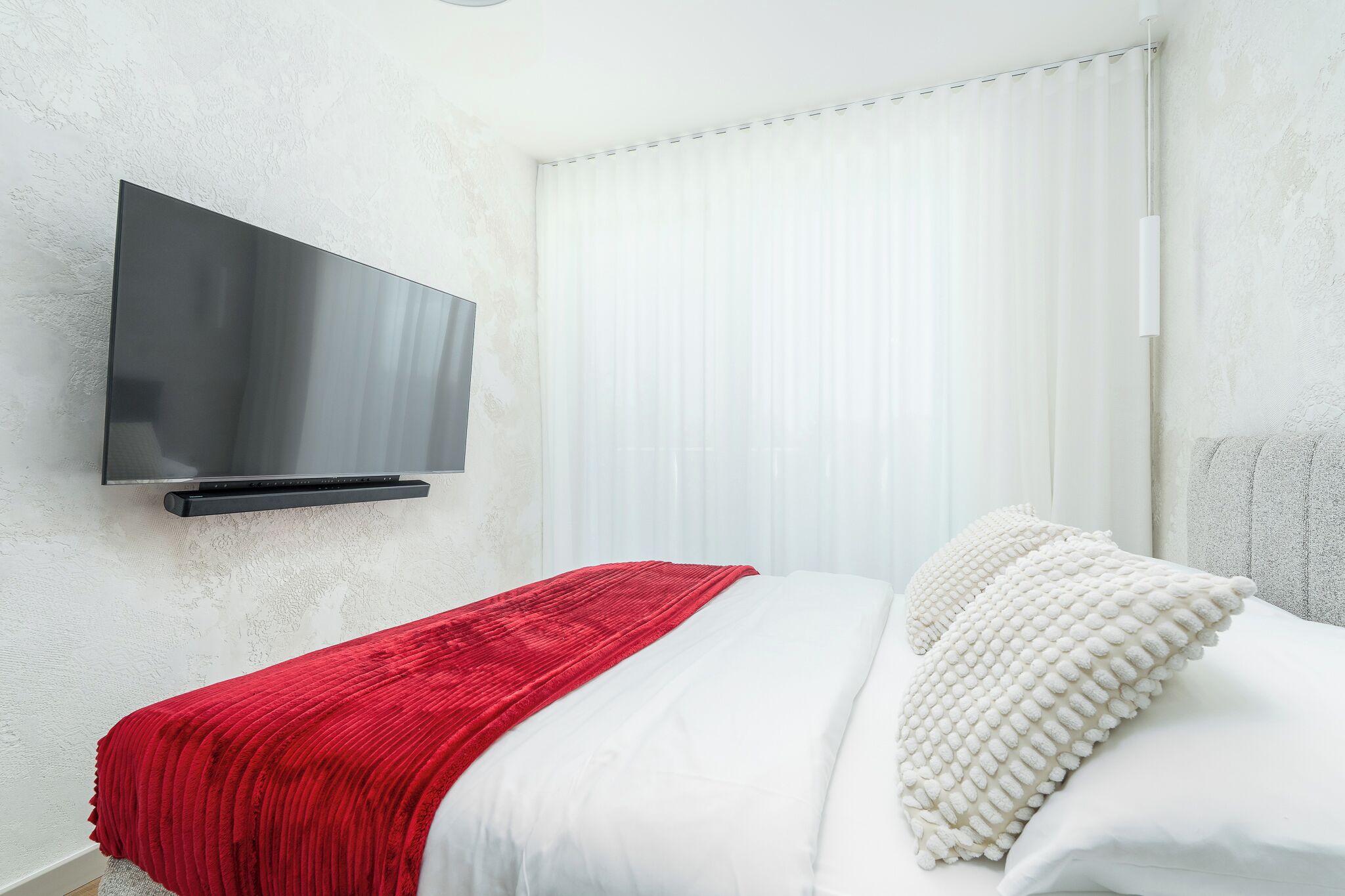 Una moderna camera d'hotel con una TV a schermo piatto fissata al muro, un letto con biancheria bianca e una coperta rossa, e un'ampia finestra con tende bianche.