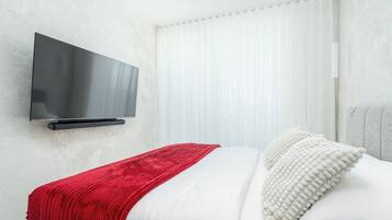 Una moderna camera d'hotel con una TV a schermo piatto fissata al muro, un letto con biancheria bianca e una coperta rossa, e un'ampia finestra con tende bianche.