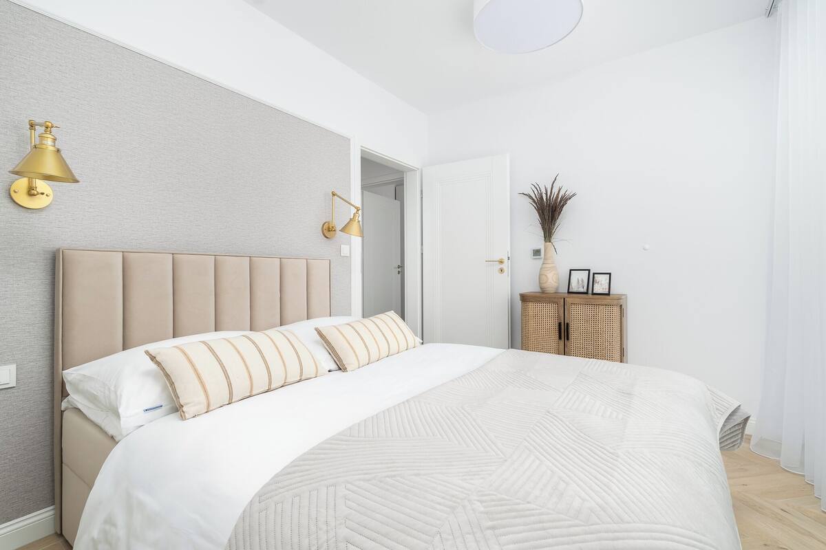 Un dormitorio con una cama grande, cabecera, dos lámparas de pared, mesita de noche con un jarrón y marcos de fotos, y una puerta que da a otra habitación.