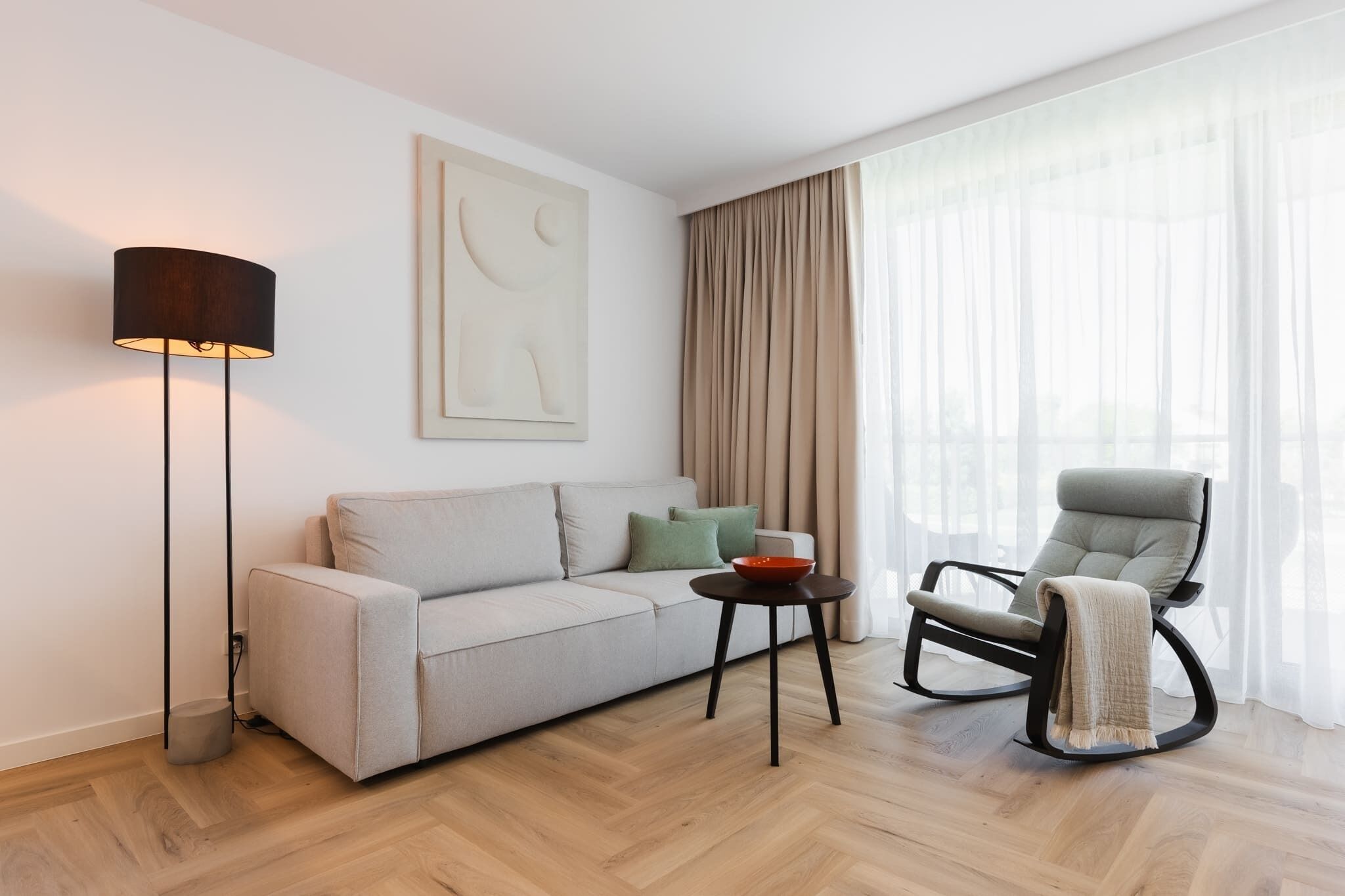 A modern living room with a sofa, a rocking chair, a floor lamp, and a painting on the wall.
