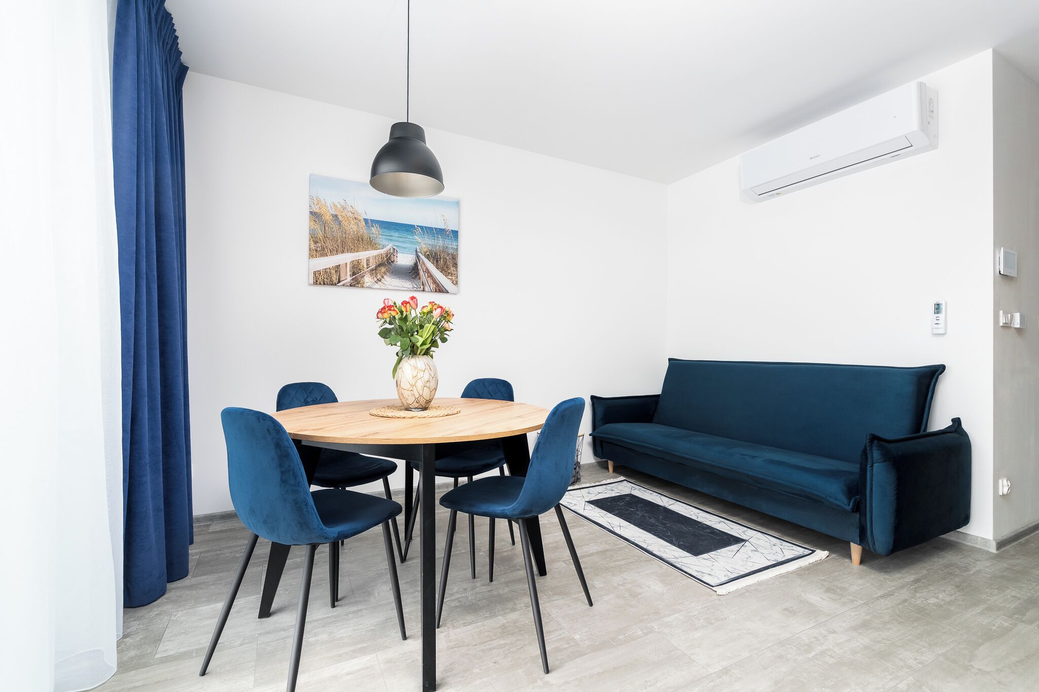 Una sala moderna con un sofá azul, una mesa redonda de comedor con sillas, una lámpara colgante y un aire acondicionado instalado en la pared.