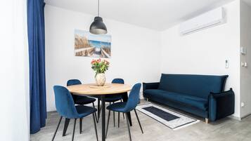 Una sala moderna con un sofá azul, una mesa redonda de comedor con sillas, una lámpara colgante y un aire acondicionado instalado en la pared.