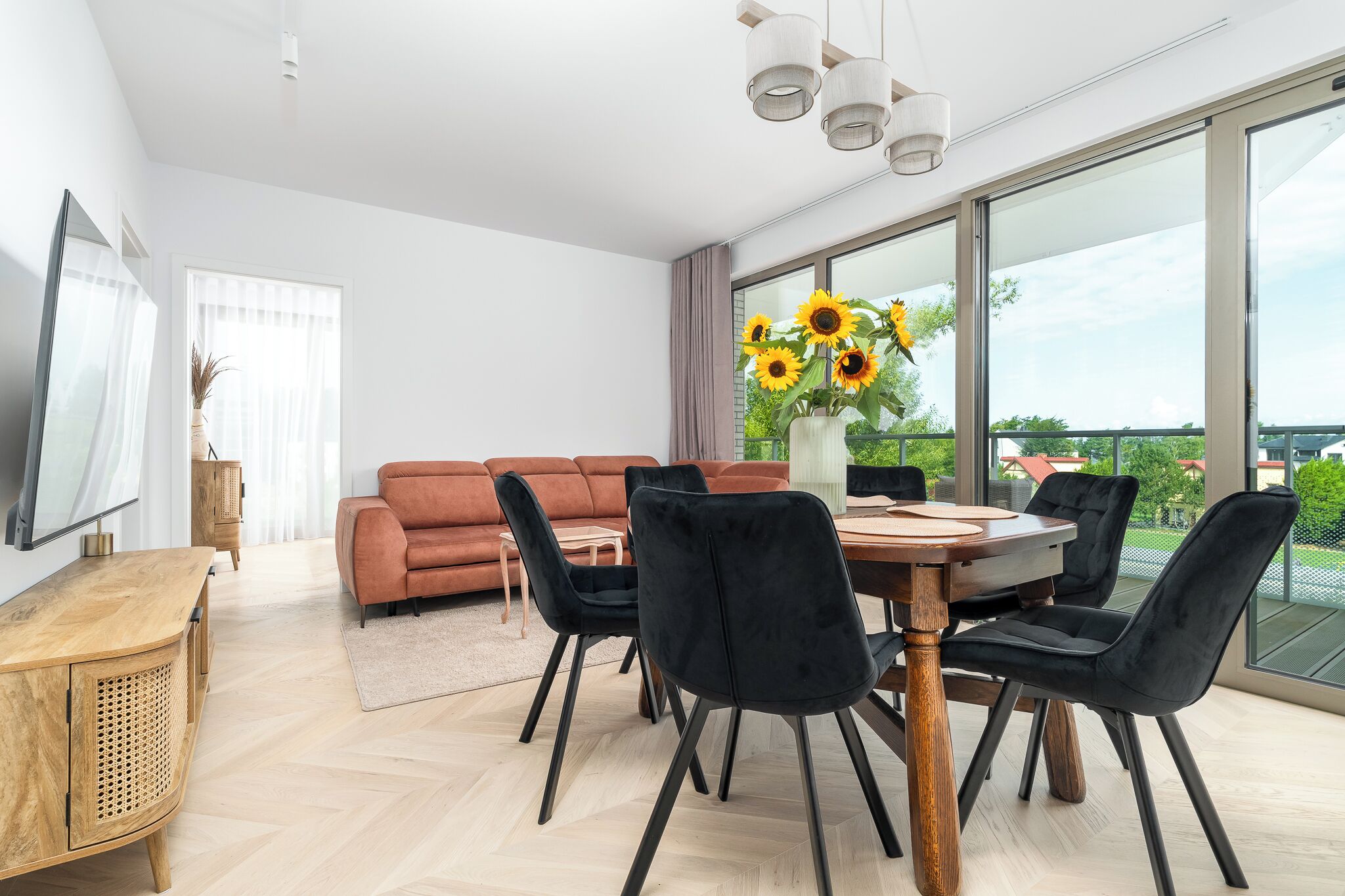 A modern living room with a wooden dining table, black chairs, a brown sofa, and a large window with a view of greenery.