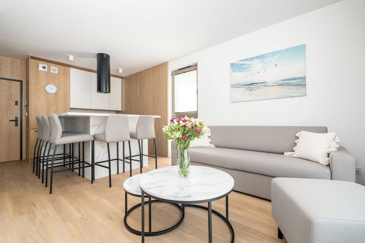 Una sala de estar moderna con un sofá gris, una mesa de centro redonda de mármol y una zona de cocina con barra y taburetes.