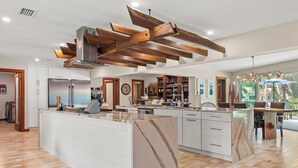 A modern kitchen with a large island, wooden ceiling beams, and a view of the outdoors. - Rustic Sands - 650 “Siesta Key” (Sarasota)