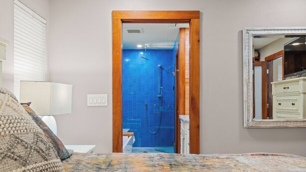 A bedroom with a bed, a nightstand, a lamp, a wooden door leading to a bathroom with blue tiles, and a mirror on the wall. - Rustic Sands - 650 “Siesta Key” (Sarasota)