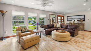 A spacious living room with a brown leather sofa, a beige armchair, and a round ottoman. There is a ceiling fan, a large window with blinds, and a flat-screen TV mounted on the wall. - Rustic Sands - 650   “Siesta Key” (Sarasota)