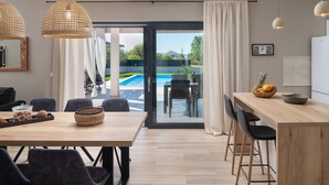 Eine moderne Küche mit einer Holzauslage, ein Essbereich mit Tisch und Stühlen und Blick auf einen Swimmingpool durch Glastüren.