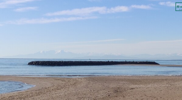 Une plage avec une digue rocheuse qui s’avance dans des eaux calmes, un rivage sablonneux et, au loin, des montagnes sous un ciel dégagé.