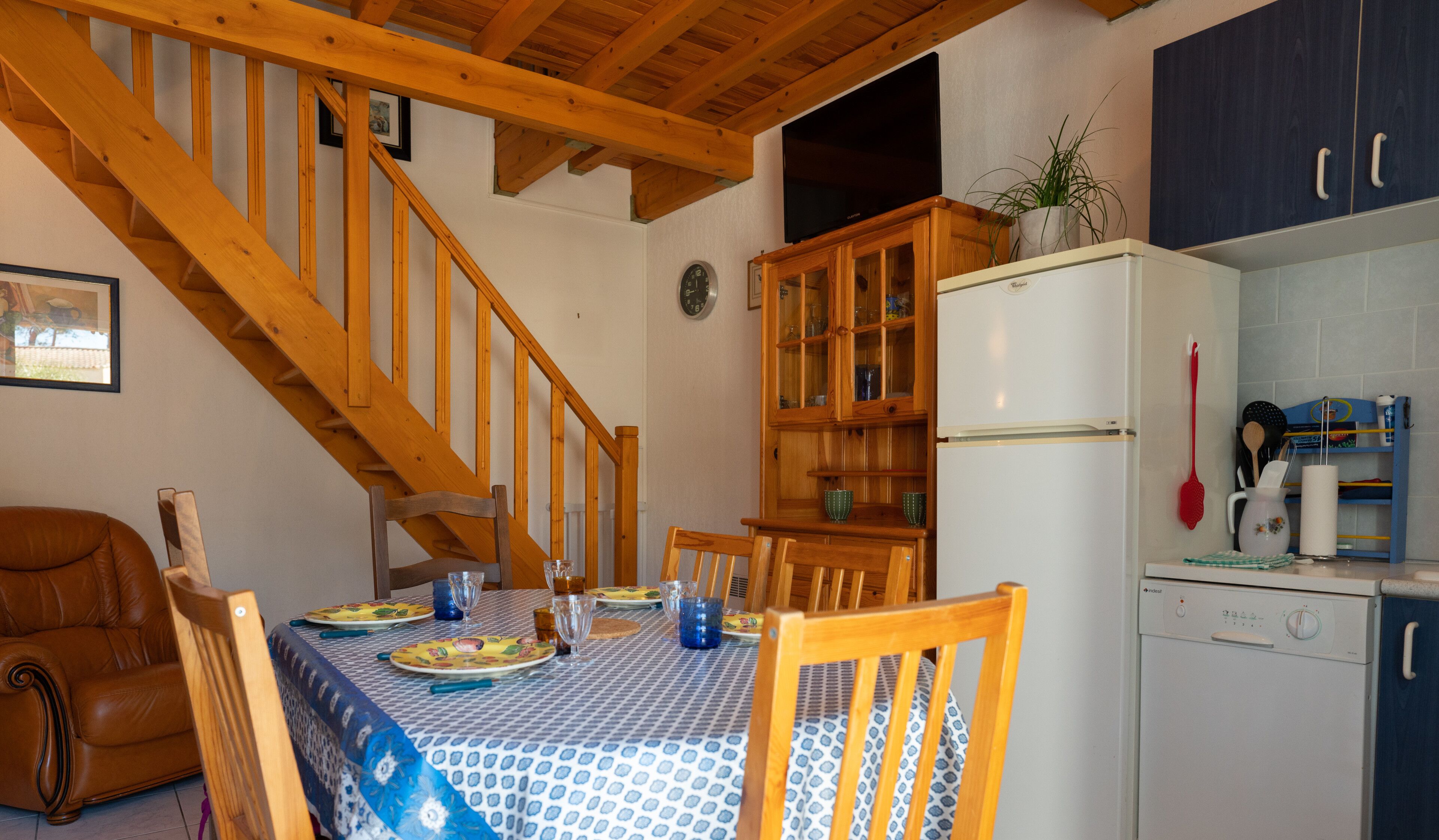 Un coin repas avec un escalier en bois, une table à manger dressée avec des assiettes et des verres, un réfrigérateur et un espace cuisine équipé de placards et d’ustensiles.