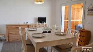 Ein Essbereich mit einem Holztisch, gedeckt für vier Personen, weißen Stühlen, einer gewebten Pendelleuchte und einem Holzschrank mit einem Flachbildfernseher.