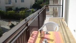 Un balcone con un tavolo apparecchiato per due, una sedia e un contenitore in metallo, con vista su una strada e degli edifici.