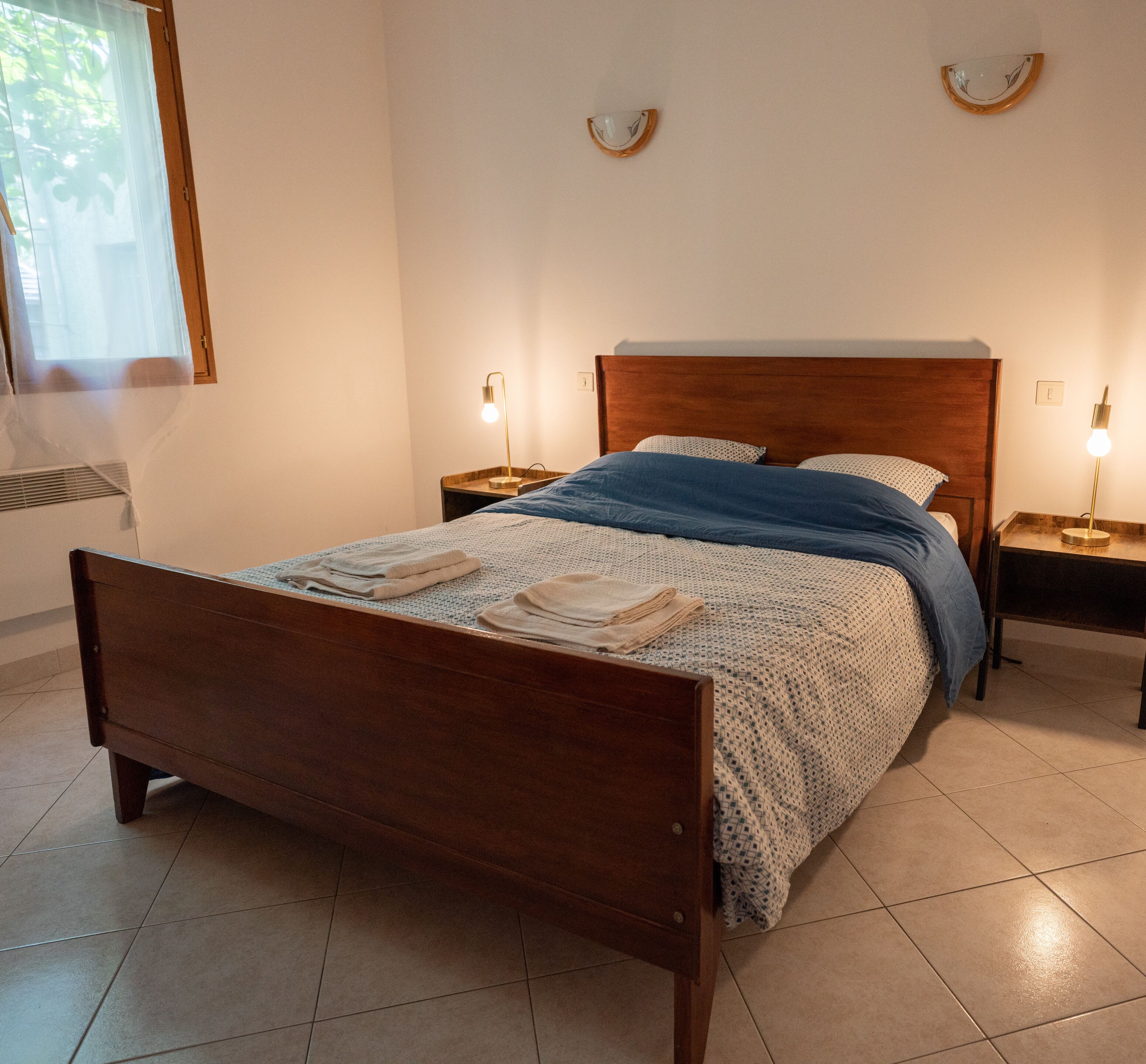 Un lit bien fait, avec une tête de lit en bois, deux tables de chevet avec des lampes et une fenêtre ornée de rideaux légers.