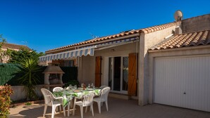 A patio with a table set for four, surrounded by white chairs, under a covered area with a striped awning.