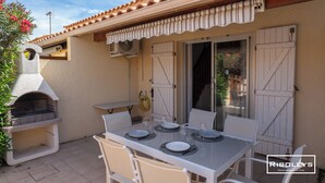Eine Terrasse im Freien mit Esstisch, Grill und einer Glastür zu einem überdachten Bereich.