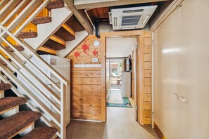 Un escalier en bois avec une rampe, un climatiseur fixé au plafond et une porte donnant sur une pièce avec un lit et une petite table.
