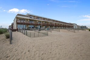 Un hotel frente a la playa con varios balcones, una playa arenosa en primer plano y cielo despejado.
