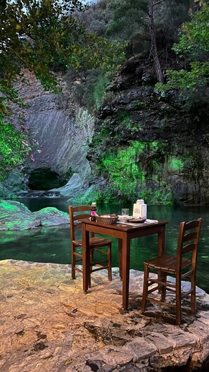A table and two chairs set on a rocky ledge overlooking a river with a cave in the background. - Kazdağı Atilabey Konağı Oteli (Ayvacik)
