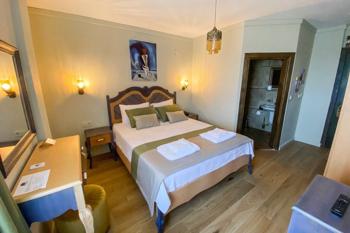 A hotel room with a large bed, bedside tables, a desk, a chair, a mirror, a painting on the wall, and a bathroom visible through an open door.
