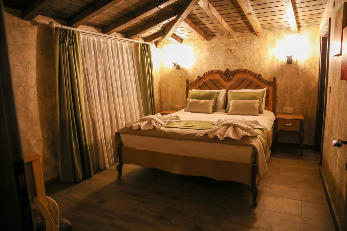 A bedroom with a wooden bed, a nightstand, a wall-mounted lamp, and a window with curtains.