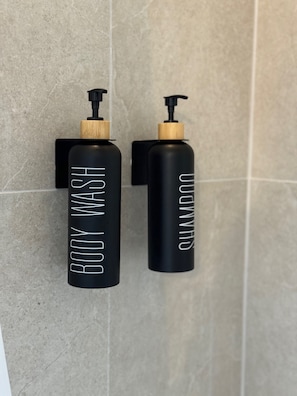 Two black bottles with pumps and wooden caps, labeled "BODY WASH" and "SHAMPOO," mounted on a tiled wall.