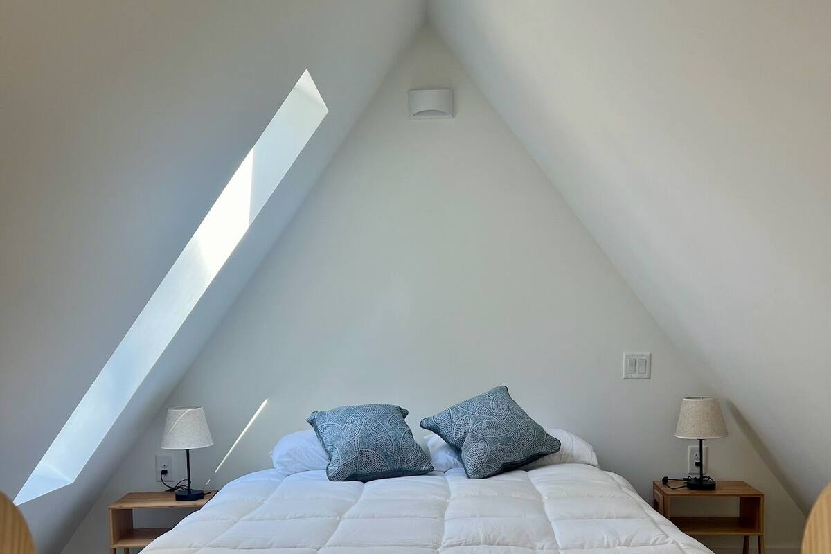 A bedroom with a sloped ceiling, a large bed with a white duvet, two matching pillows, and two bedside tables with lamps.