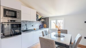 Una cocina moderna con armarios blancos, horno empotrado y microondas. Hay una mesa de comedor con sillas y un jarrón con ramas.