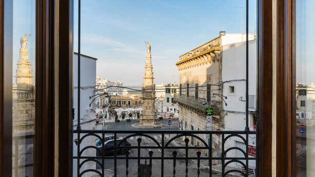 A view from a window overlooking a city square with historic buildings and a monument.