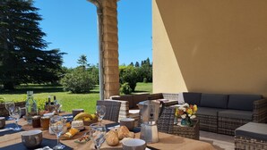 Ein Essbereich im Freien mit Tisch, der für eine Mahlzeit gedeckt ist, Korbmöbel und Blick auf den Garten.