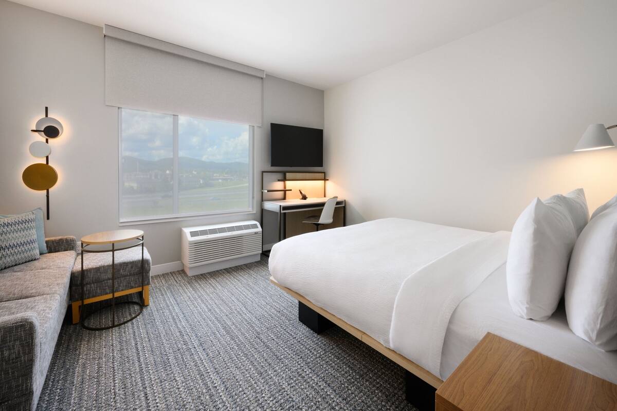 Une chambre d’hôtel moderne dotée d’un grand lit, d’un bureau, d’une chaise, d’une petite table, d’une télévision et d’une fenêtre donnant sur un beau paysage.