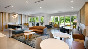 A spacious, well-lit living area with a sofa, coffee table, and dining table. Large windows provide natural light and a view of greenery outside.