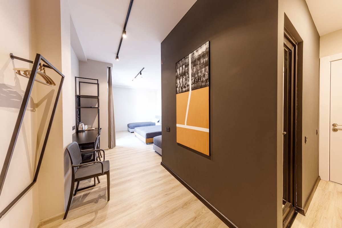 A modern hotel corridor with a dark wall, a framed artwork, a chair, a table, and a bed in the background.