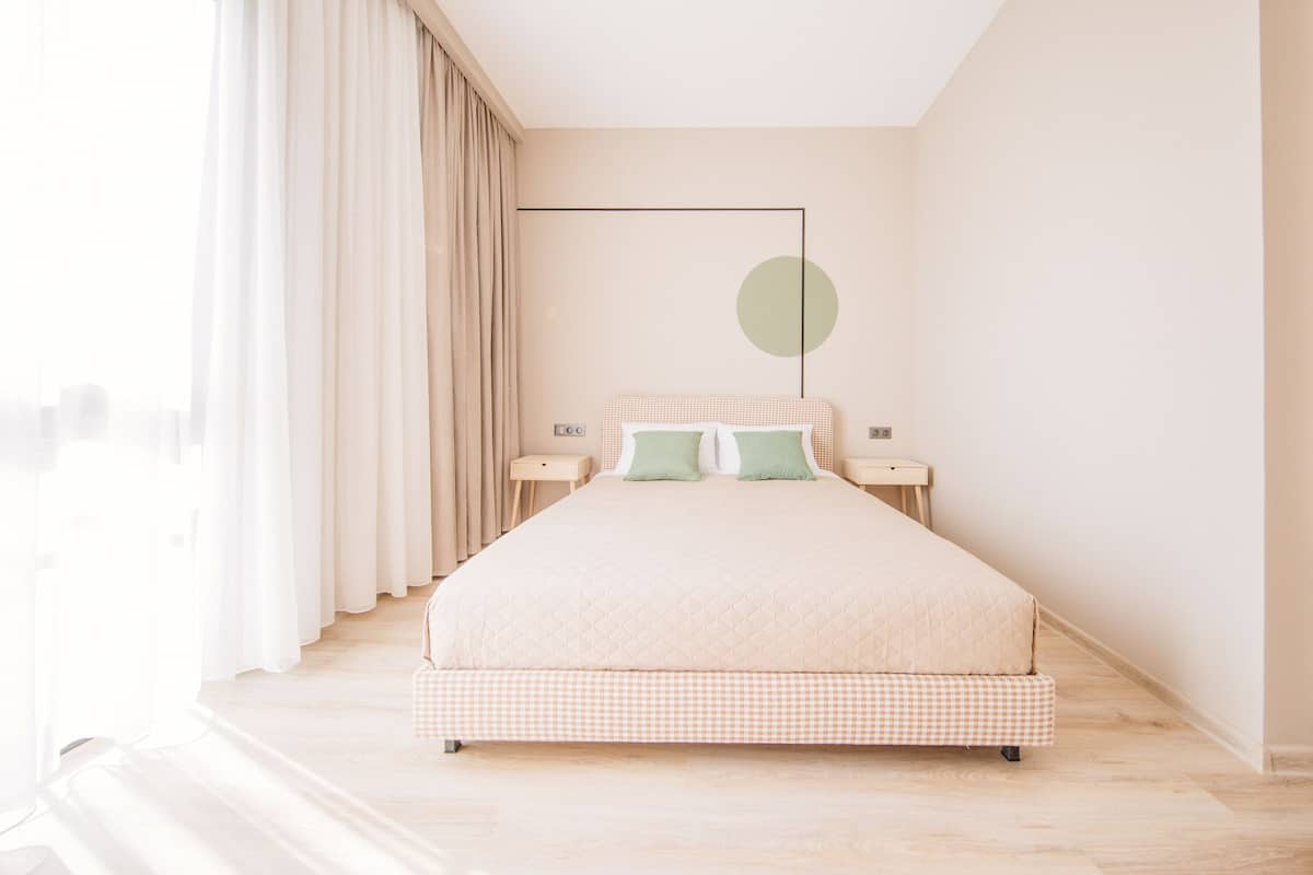 A bedroom with a large bed, two bedside tables, a hanging light fixture, and a view of the city through sheer curtains.