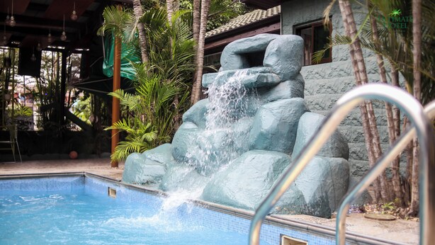 A swimming pool with a waterfall feature, surrounded by tropical plants and a stone wall. - ULTIMATE HOTEL (Mile 11)