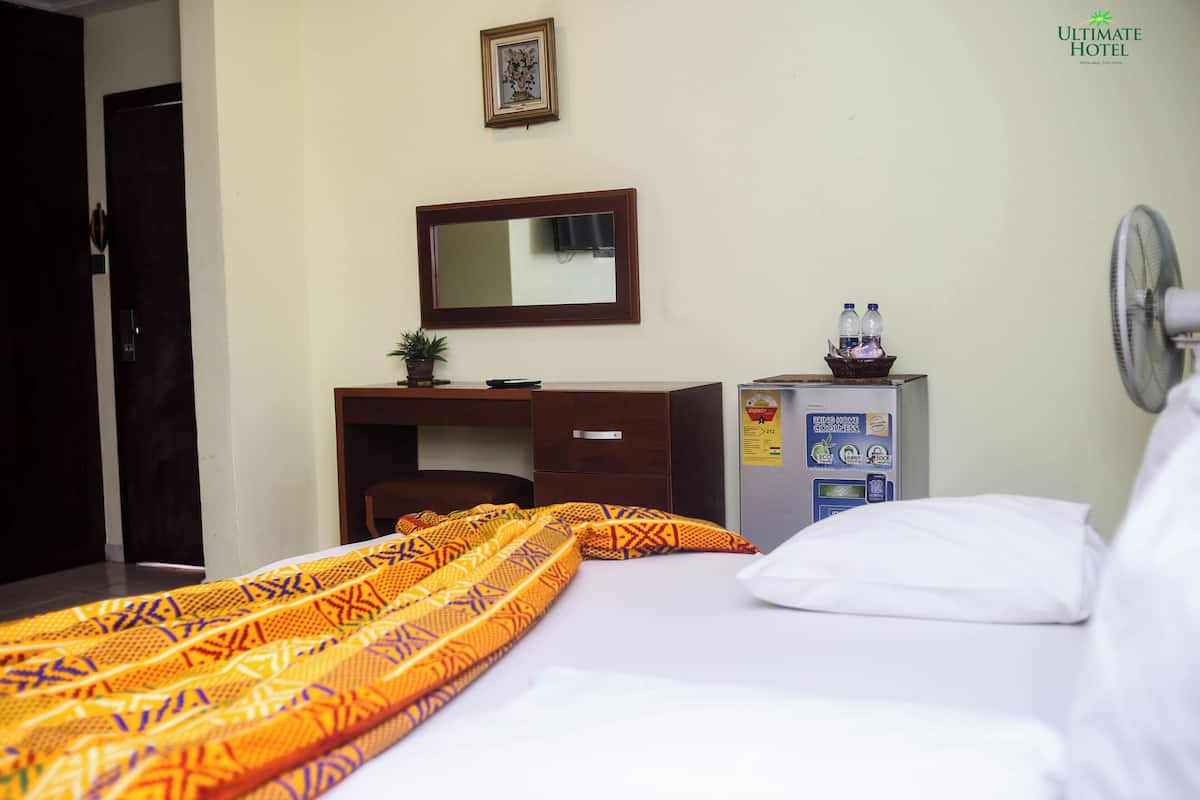 Una habitación de hotel con cama, escritorio, silla, espejo, ventilador de pared y máquina expendedora.