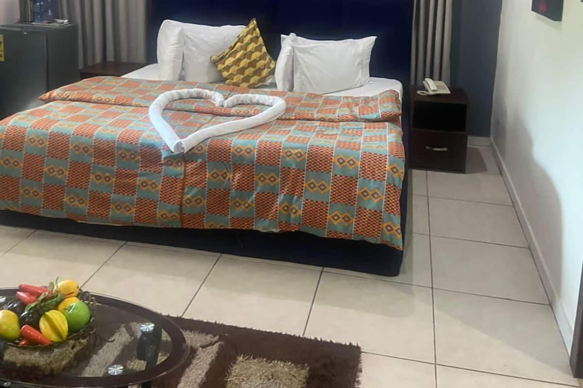 Habitación de hotel con cama, cortinas, equipo de aire acondicionado, mesita de noche con teléfono y una pequeña mesa con fruta.
