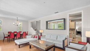 A modern living room with a sofa, coffee table, dining table, and chairs. A staircase is visible in the background.