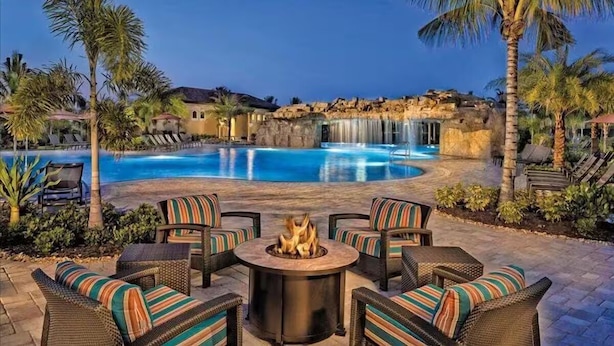 Zona de piscina con un hogar y sillones reclinables, rodeada de palmeras y una cascada.