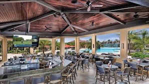 Zona de bar al aire libre con techo, múltiples opciones de asientos, vista a la piscina y una pantalla de televisión.