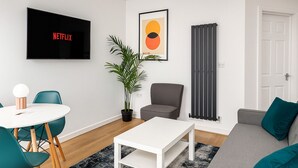 A modern living room with a flat-screen TV, a dining table with chairs, a grey sofa, a potted plant, and a radiator. - Perfect Modern 4 bedroom House in Liverpool with Garden - Free Wi-Fi - Parking (Tuebrook)