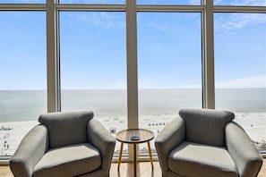 Een kamer met een groot raam met uitzicht op een strand, twee fauteuils en een kleine ronde tafel.