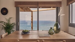 A dining area with a wooden table, chairs, a bowl of fruit, a vase, and a view of the sea through large windows.