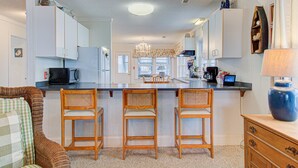 A kitchen with white cabinets, a central island with bar stools, a chandelier, and a dining area with a wicker sofa. - ⛱️The Beach House-Cozy Cottage w/ Ocean views!⛱️ (Carolina Beach)